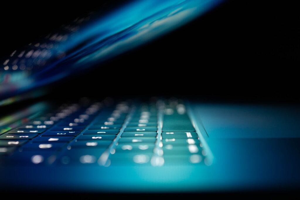 A close-up of a laptop keyboard illuminated in blue light, with a blurred screen in the background.